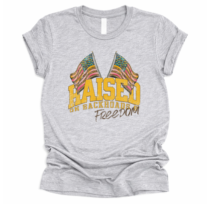 Patriotic Americana graphic on a t-shirt. The design features bold varsity-style “RAISED” lettering with crossed American flags above, finished with “on backroads” and “Freedom” in script below. The distressed, vintage-inspired texture and warm tones make it an easy Fourth of July t-shirt.  gray color tee
