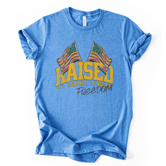 Patriotic Americana graphic on a t-shirt. The design features bold varsity-style “RAISED” lettering with crossed American flags above, finished with “on backroads” and “Freedom” in script below. The distressed, vintage-inspired texture and warm tones make it an easy Fourth of July t-shirt.  blue color tee