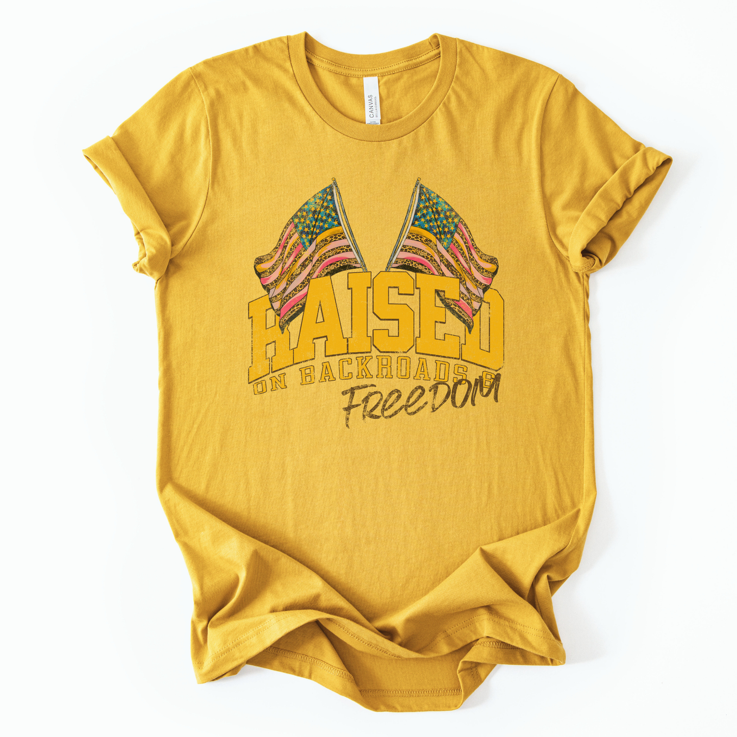 Patriotic Americana graphic on a t-shirt. The design features bold varsity-style “RAISED” lettering with crossed American flags above, finished with “on backroads” and “Freedom” in script below. The distressed, vintage-inspired texture and warm tones make it an easy Fourth of July t-shirt.  yellow color tee