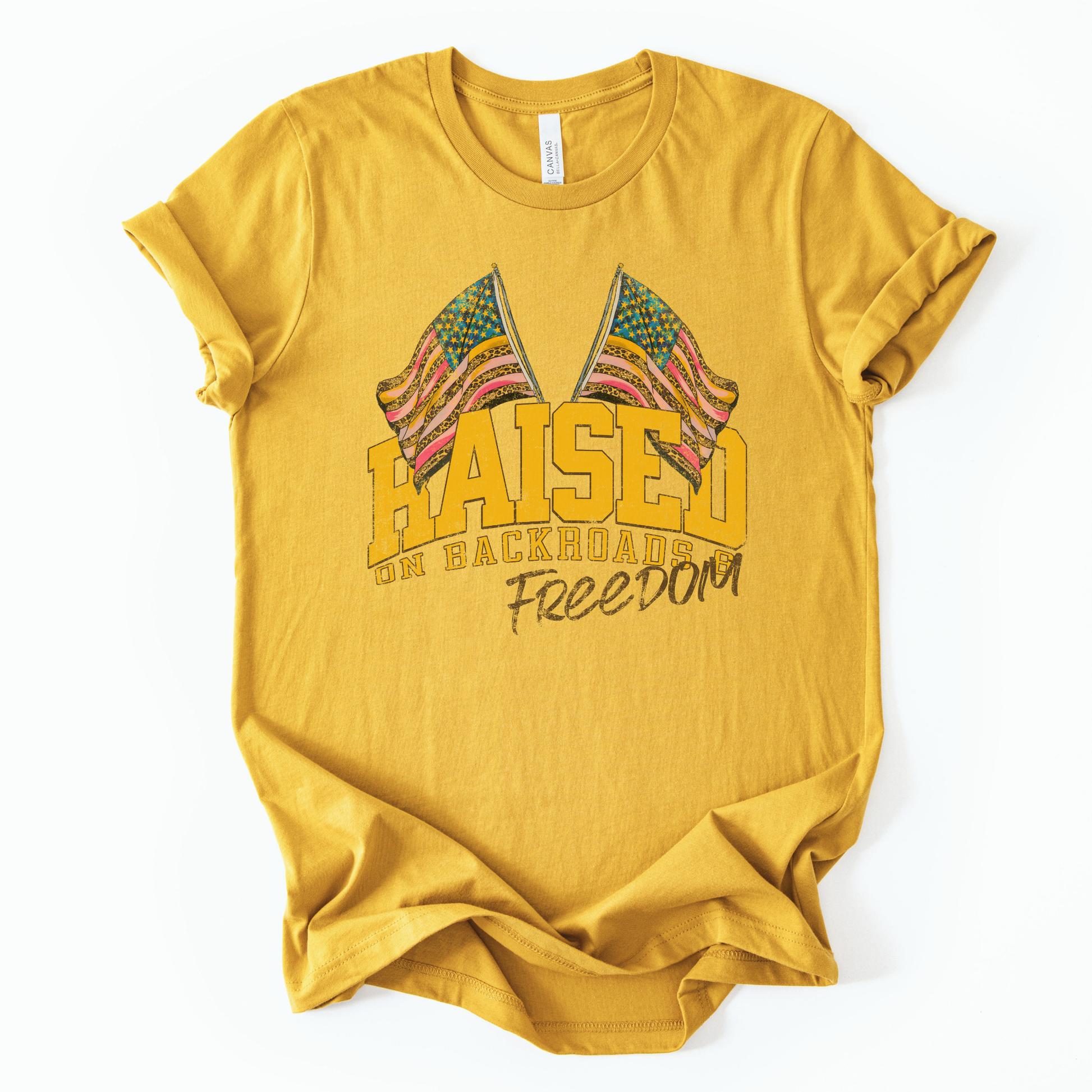 Patriotic Americana graphic on a t-shirt. The design features bold varsity-style “RAISED” lettering with crossed American flags above, finished with “on backroads” and “Freedom” in script below. The distressed, vintage-inspired texture and warm tones make it an easy Fourth of July t-shirt.  yellow color tee