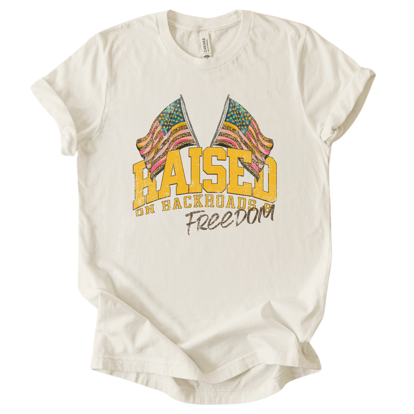 Patriotic Americana graphic on a t-shirt. The design features bold varsity-style “RAISED” lettering with crossed American flags above, finished with “on backroads” and “Freedom” in script below. The distressed, vintage-inspired texture and warm tones make it an easy Fourth of July t-shirt.  Cream color tee