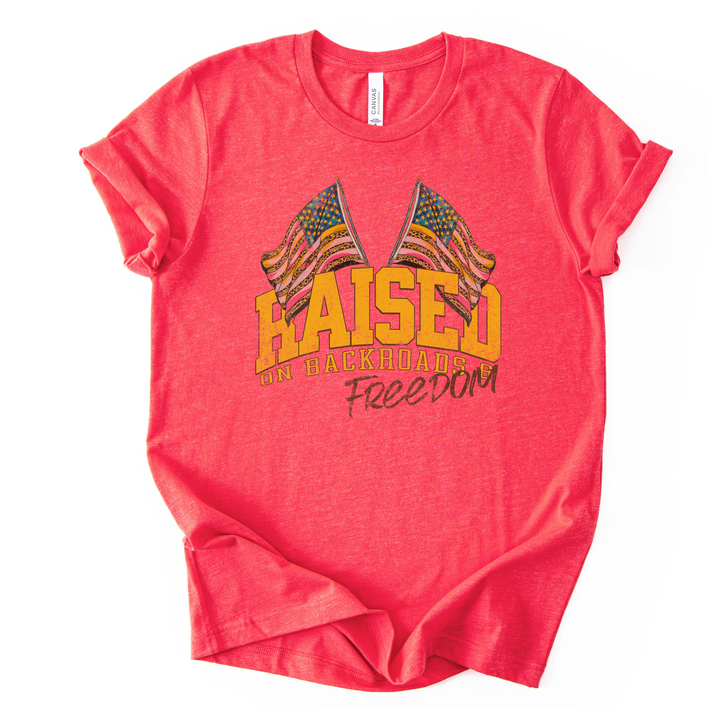 Patriotic Americana graphic on a t-shirt. The design features bold varsity-style “RAISED” lettering with crossed American flags above, finished with “on backroads” and “Freedom” in script below. The distressed, vintage-inspired texture and warm tones make it an easy Fourth of July t-shirt.  red color tee