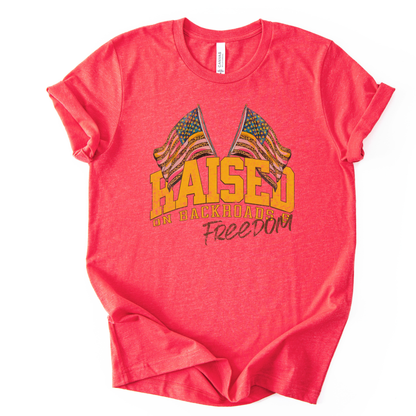 Patriotic Americana graphic on a t-shirt. The design features bold varsity-style “RAISED” lettering with crossed American flags above, finished with “on backroads” and “Freedom” in script below. The distressed, vintage-inspired texture and warm tones make it an easy Fourth of July t-shirt.  red color tee