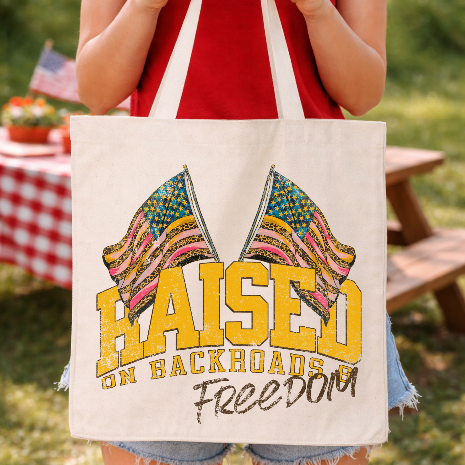 Patriotic Fourth of July country-inspired graphic on a 100% cotton canvas tote bag. The design features bold “Raised on Backroads” lettering with crossed American flags filled with mixed stars, stripes, and pattern accents, finished with a distressed “Freedom” script for a rustic Americana look that resonates with rural and small-town shoppers.