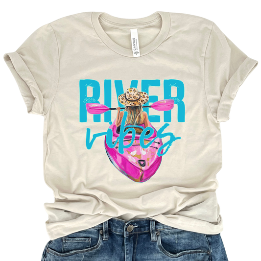 River Vibes kayak graphic on a soft unisex Bella Canvas heather oatmeal t shirt. The artwork shows a woman paddling a kayak from a back view wearing a leopard print sun hat, with pink paddles stretching across the top of the design.
