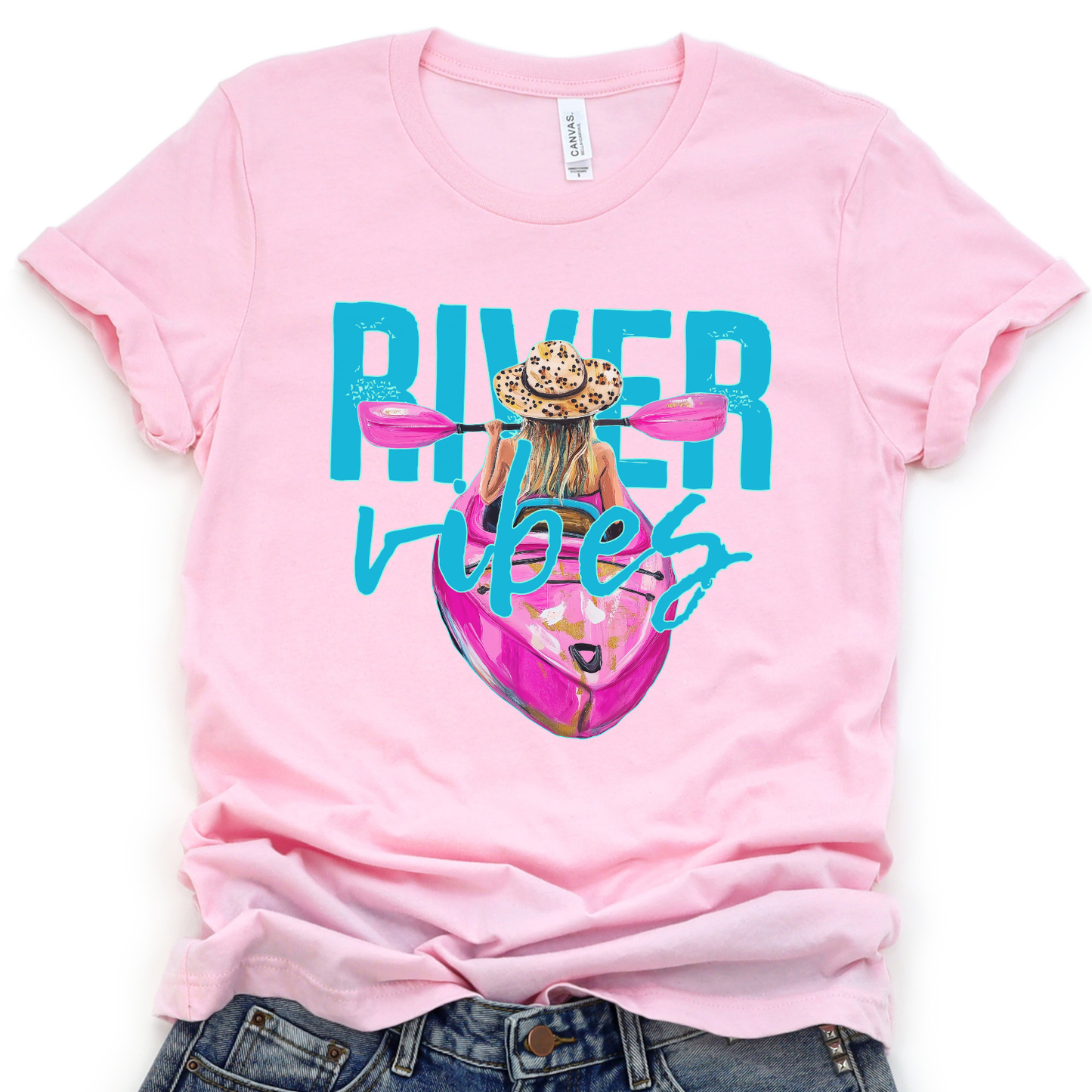 River Vibes kayak graphic on a soft unisex Bella Canvas heather pink t shirt. The artwork shows a woman paddling a kayak from a back view wearing a leopard print sun hat, with pink paddles stretching across the top of the design.