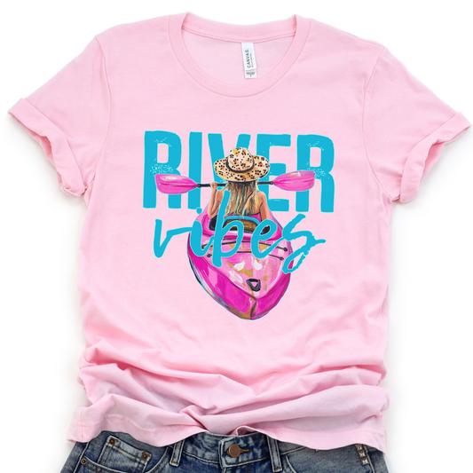 River Vibes kayak graphic on a soft unisex Bella Canvas heather pink t shirt. The artwork shows a woman paddling a kayak from a back view wearing a leopard print sun hat, with pink paddles stretching across the top of the design.