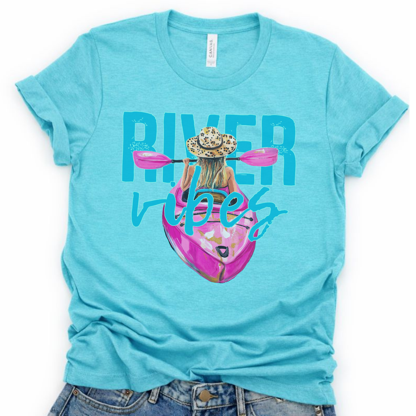 River Vibes kayak graphic on a soft unisex Bella Canvas heather aqua t shirt. The artwork shows a woman paddling a kayak from a back view wearing a leopard print sun hat, with pink paddles stretching across the top of the design.