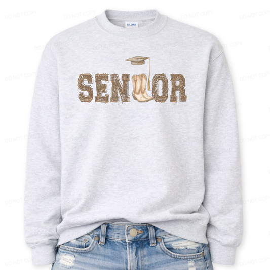 Western graduation graphic on a classic ash crewneck sweatshirt. The design features SENIOR in bold varsity style lettering with a pair of cowboy boots integrated into the wordmark and a small graduation cap detail above,
