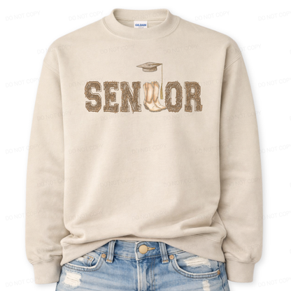 Western graduation graphic on a classic sand crewneck sweatshirt. The design features SENIOR in bold varsity style lettering with a pair of cowboy boots integrated into the wordmark and a small graduation cap detail above,