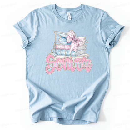 Graduation 2026 academic themed graphic t shirt. The design features a stacked book illustration topped with a light blue graduation cap and tassel, accented with a large pink bow. The front reads Senior with Twenty Twenty Six layered underneath, creating a preppy graduation look 