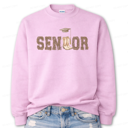 Western graduation graphic on a classic pink crewneck sweatshirt. The design features SENIOR in bold varsity style lettering with a pair of cowboy boots integrated into the wordmark and a small graduation cap detail above,