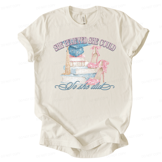 Graduation themed inspirational graphic t shirt. The design features a stacked book illustration topped with a blue graduation cap and long tassel, paired with pink high heel shoes tied with bows. The artwork reads She Believed She Could across the top and So She Did in script below, creating a soft academic graduation look with strong gift appeal.