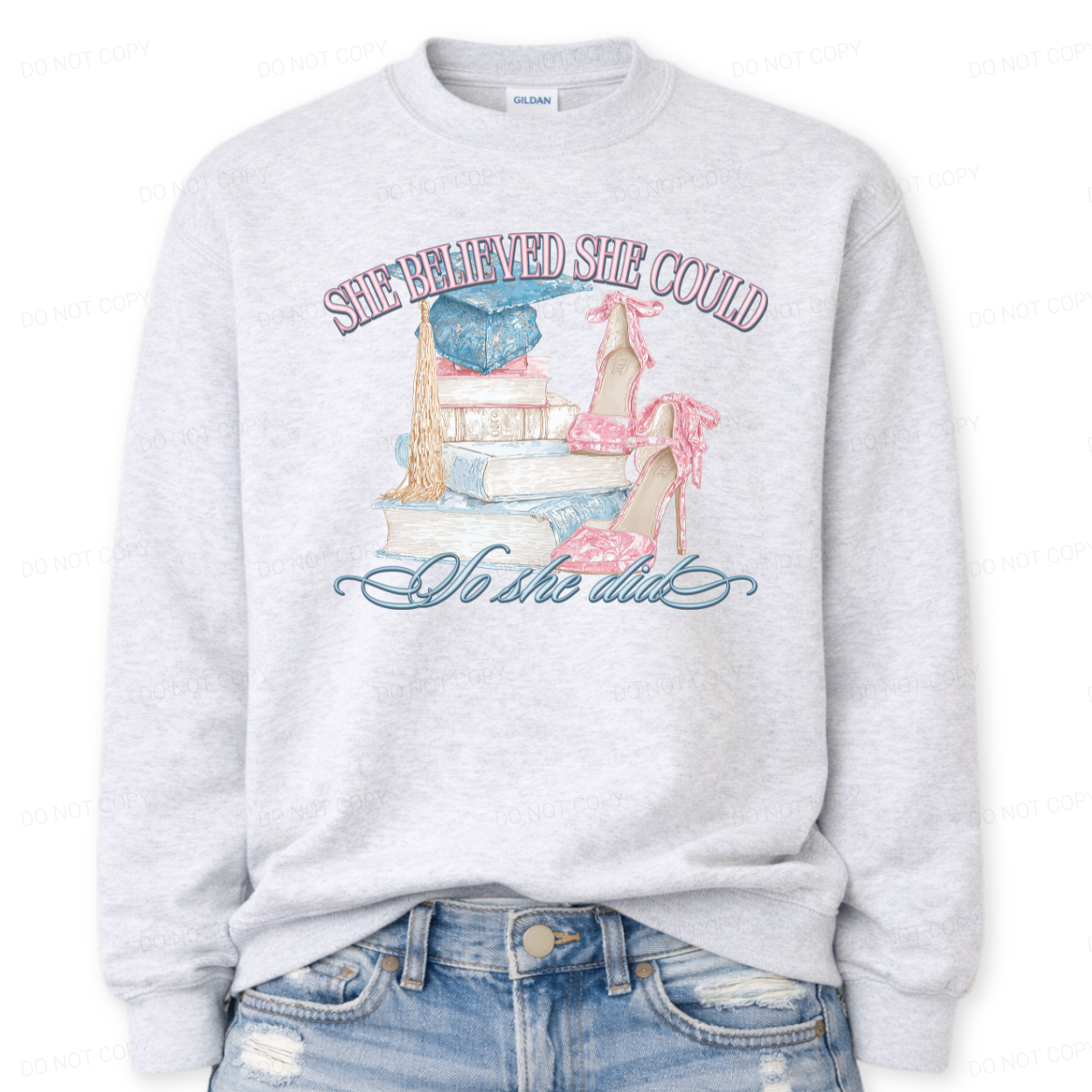Inspirational graduation graphic on a classic ash crewneck sweatshirt. The design features the phrase She Believed She Could So She Did arched above and below a detailed illustration of stacked books, a blue graduation cap with gold tassel, and pink toile high heels. The soft pastel tones and academic elements create a feminine graduation look that works for both high school seniors and college graduates.