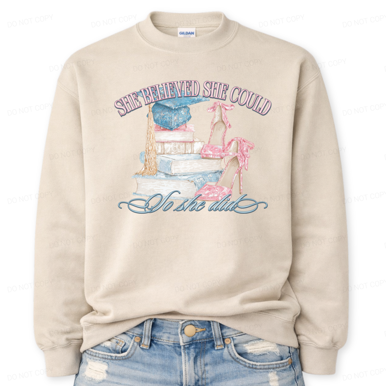 Inspirational graduation graphic on a classic crewneck sweatshirt. The design features the phrase She Believed She Could So She Did arched above and below a detailed illustration of stacked books, a blue graduation cap with gold tassel, and pink toile high heels. The soft pastel tones and academic elements create a feminine graduation look that works for both high school seniors and college graduates.