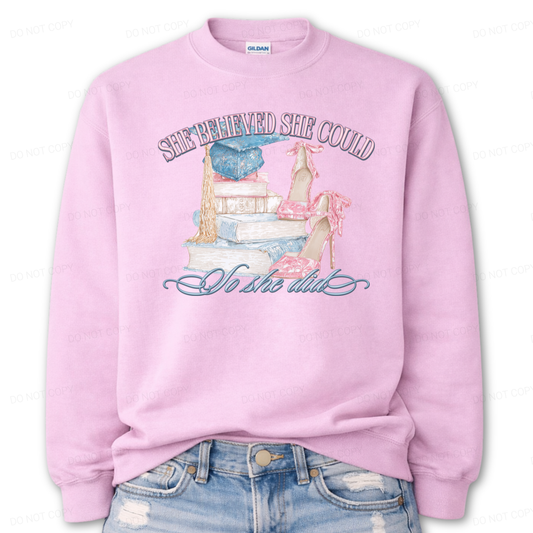 Inspirational graduation graphic on a classic crewneck sweatshirt. The design features the phrase She Believed She Could So She Did arched above and below a detailed illustration of stacked books, a blue graduation cap with gold tassel, and pink toile high heels. The soft pastel tones and academic elements create a feminine graduation look that works for both high school seniors and college graduates.