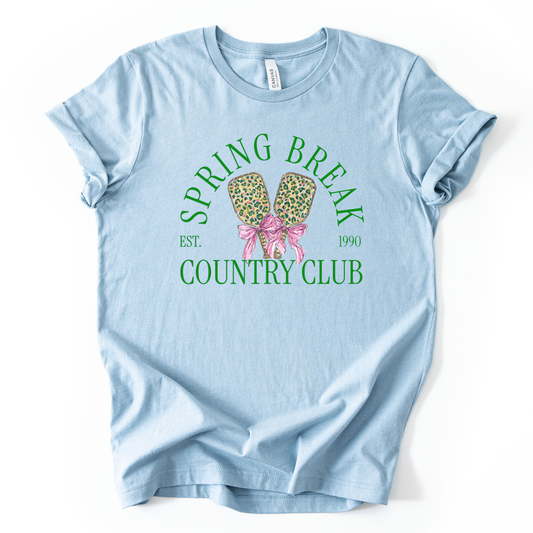blue t-shirt with 'Spring Break Country Club' graphic on a white background.The trending graphic features a pair of illustrated pickleball paddles with a leopard-style pattern, tied together with a pink ribbon bow at the center. Arched text reads “Spring Break Country Club”, with small accent text adding a vintage-inspired touch. The overall design feels playful, preppy, and spring-ready.