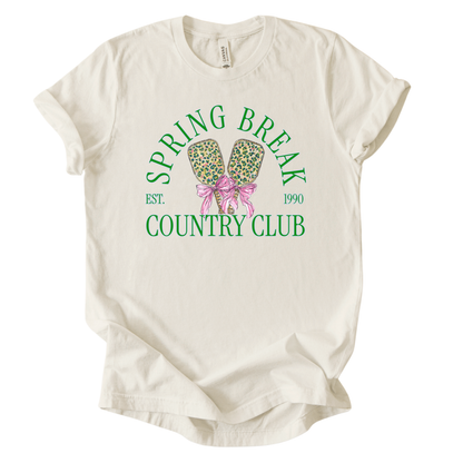 ivory t-shirt with 'Spring Break Country Club' graphic on a white background.The trending graphic features a pair of illustrated pickleball paddles with a leopard-style pattern, tied together with a pink ribbon bow at the center. Arched text reads “Spring Break Country Club”, with small accent text adding a vintage-inspired touch. The overall design feels playful, preppy, and spring-ready.