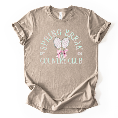 tan t-shirt with 'Spring Break Country Club' text and graphics on a white background.The graphic features a pair of illustrated tennis rackets crossed at the handles and tied together with a soft pink ribbon bow at the center. Arched text reads “Spring Break Country Club”, with small accent details adding a classic, collegiate feel. 