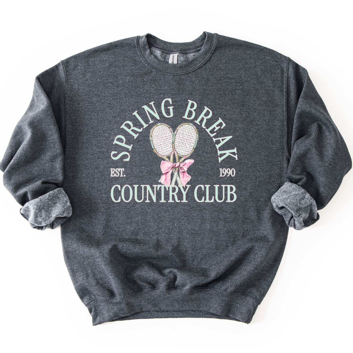 Gray Preppy spring tennis country club graphic on a sweatshirt. The graphic features the phrase “Spring Break Country Club” in arched collegiate lettering, with small “EST.” and “1990” accents on each side. Two illustrated tennis racquets are centered beneath the text and tied together with a soft pink bow, creating a polished yet playful design that blends sporty charm with spring color appeal. 
