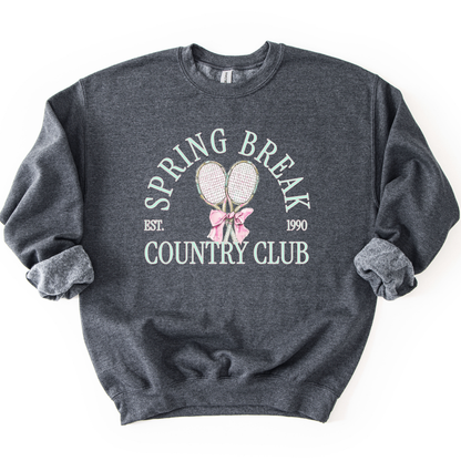 Gray Preppy spring tennis country club graphic on a sweatshirt. The graphic features the phrase “Spring Break Country Club” in arched collegiate lettering, with small “EST.” and “1990” accents on each side. Two illustrated tennis racquets are centered beneath the text and tied together with a soft pink bow, creating a polished yet playful design that blends sporty charm with spring color appeal. 