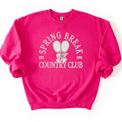 Pink Preppy spring tennis country club graphic on a sweatshirt. The graphic features the phrase “Spring Break Country Club” in arched collegiate lettering, with small “EST.” and “1990” accents on each side. Two illustrated tennis racquets are centered beneath the text and tied together with a soft pink bow, creating a polished yet playful design that blends sporty charm with spring color appeal. 