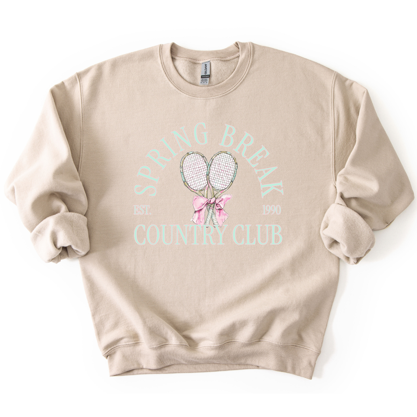 Sand Preppy spring tennis country club graphic on a sweatshirt. The graphic features the phrase “Spring Break Country Club” in arched collegiate lettering, with small “EST.” and “1990” accents on each side. Two illustrated tennis racquets are centered beneath the text and tied together with a soft pink bow, creating a polished yet playful design that blends sporty charm with spring color appeal. 