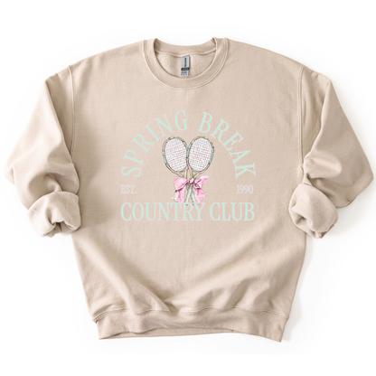 Sand Preppy spring tennis country club graphic on a sweatshirt. The graphic features the phrase “Spring Break Country Club” in arched collegiate lettering, with small “EST.” and “1990” accents on each side. Two illustrated tennis racquets are centered beneath the text and tied together with a soft pink bow, creating a polished yet playful design that blends sporty charm with spring color appeal. 