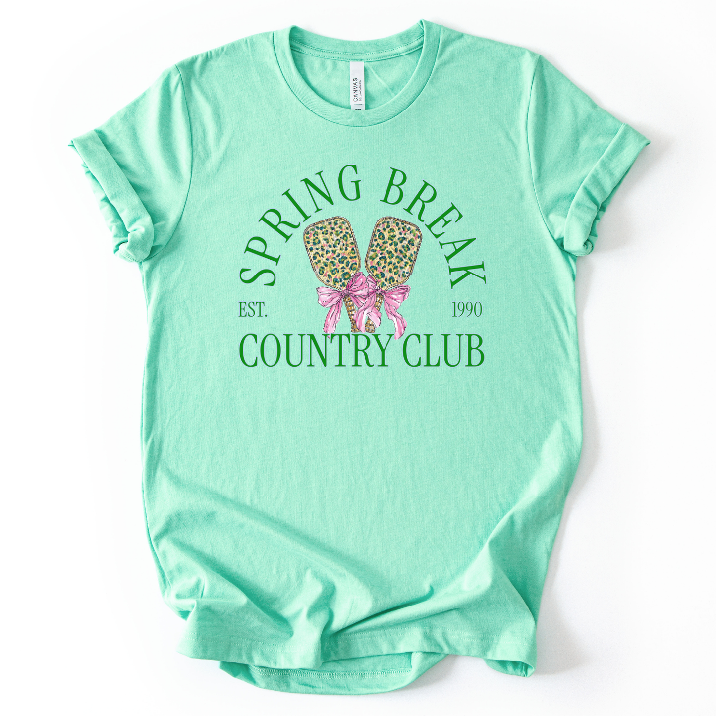 Mint green t-shirt with 'Spring Break Country Club' graphic on a white background.The trending graphic features a pair of illustrated pickleball paddles with a leopard-style pattern, tied together with a pink ribbon bow at the center. Arched text reads “Spring Break Country Club”, with small accent text adding a vintage-inspired touch. The overall design feels playful, preppy, and spring-ready.