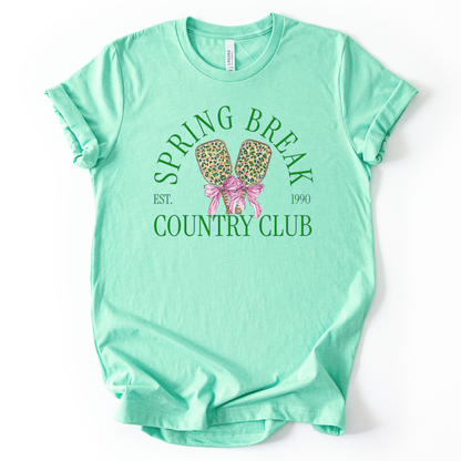 Mint green t-shirt with 'Spring Break Country Club' graphic on a white background.The trending graphic features a pair of illustrated pickleball paddles with a leopard-style pattern, tied together with a pink ribbon bow at the center. Arched text reads “Spring Break Country Club”, with small accent text adding a vintage-inspired touch. The overall design feels playful, preppy, and spring-ready.