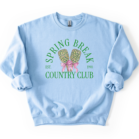 Preppy spring pickleball country club graphic on a sweatshirt. The graphic features the phrase “Spring Break Country Club” in arched collegiate lettering, with small “EST.” and “1990” accents on each side. Two illustrated pickleball paddles sit centered beneath the text and are tied together with a pink bow, creating a playful, sporty-coastal look that fits spring floor sets, pickleball gift sections, and casual lifestyle apparel displays.