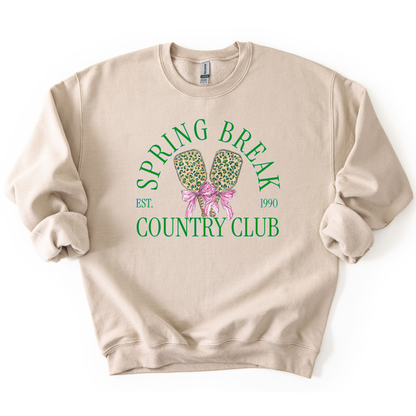 Preppy spring pickleball country club graphic on a sweatshirt. The graphic features the phrase “Spring Break Country Club” in arched collegiate lettering, with small “EST.” and “1990” accents on each side. Two illustrated pickleball paddles sit centered beneath the text and are tied together with a pink bow, creating a playful, sporty-coastal look that fits spring floor sets, pickleball gift sections, and casual lifestyle apparel displays.