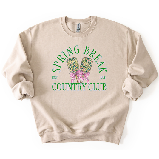 Preppy spring pickleball country club graphic on a sweatshirt. The graphic features the phrase “Spring Break Country Club” in arched collegiate lettering, with small “EST.” and “1990” accents on each side. Two illustrated pickleball paddles sit centered beneath the text and are tied together with a pink bow, creating a playful, sporty-coastal look that fits spring floor sets, pickleball gift sections, and casual lifestyle apparel displays.