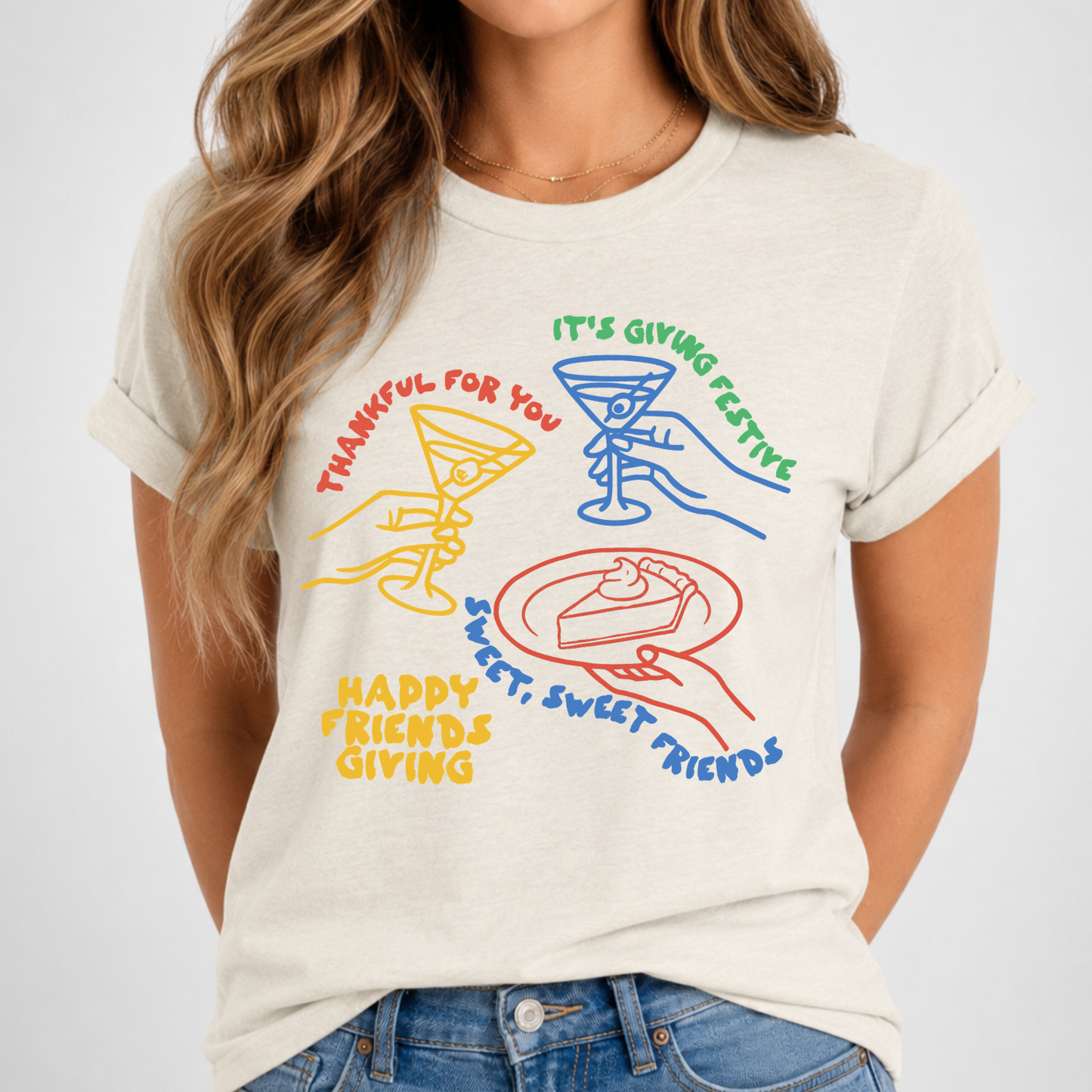 A woman wearing a light beige graphic t-shirt with colorful drawings of a martini glass, a slice of cake, and text that reads 'THANKFUL FOR YOU', 'IT'S GIVING FESTIVE', 'HAPPY FRIENDS GIVING', and 'SWEET, SWEET FRIENDS'.