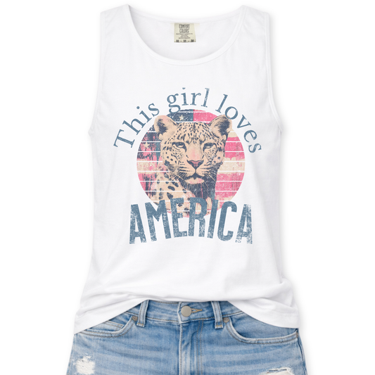 White Comfort Colors tank top featuring a vintage-style patriotic graphic with a leopard illustration inside a distressed American flag circle and the phrase “This Girl Loves America,” styled with light wash denim jeans.