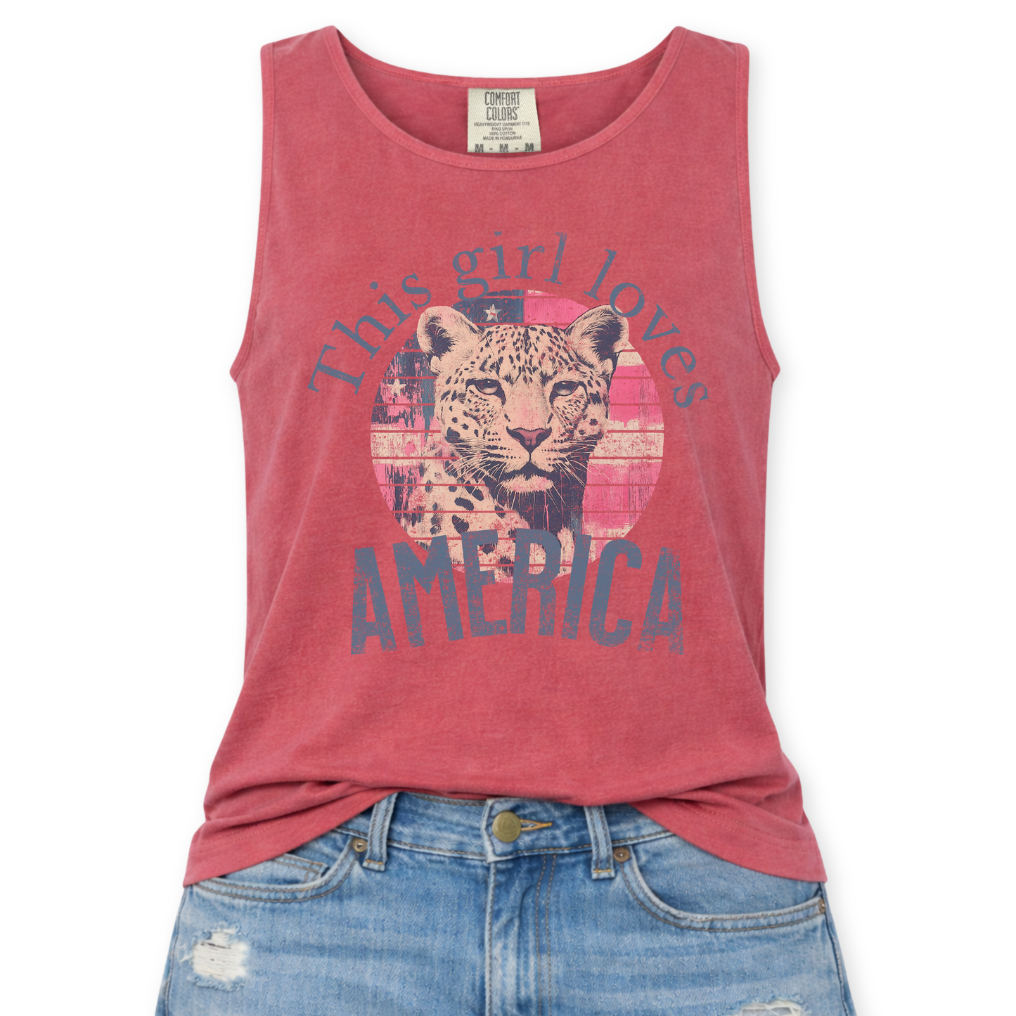 Crimson Red Comfort Colors tank top featuring a vintage-style patriotic graphic with a leopard illustration inside a distressed American flag circle and the phrase “This Girl Loves America,” styled with light wash denim jeans.