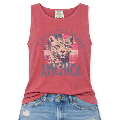Crimson Red Comfort Colors tank top featuring a vintage-style patriotic graphic with a leopard illustration inside a distressed American flag circle and the phrase “This Girl Loves America,” styled with light wash denim jeans.