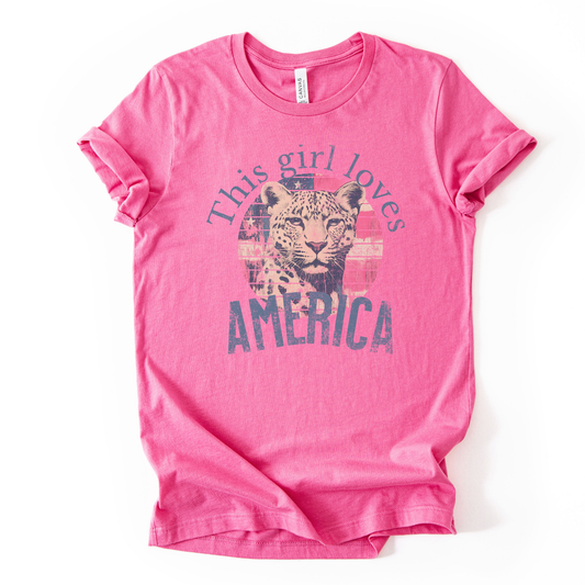 Patriotic Americana graphic on a pink t-shirt. The design features an illustrated leopard portrait centered over a distressed American flag circle, paired with the phrase “This Girl Loves America” in bold, vintage-style lettering. The worn texture and soft color palette create a modern patriotic look that blends animal imagery with classic Fourth of July appeal.