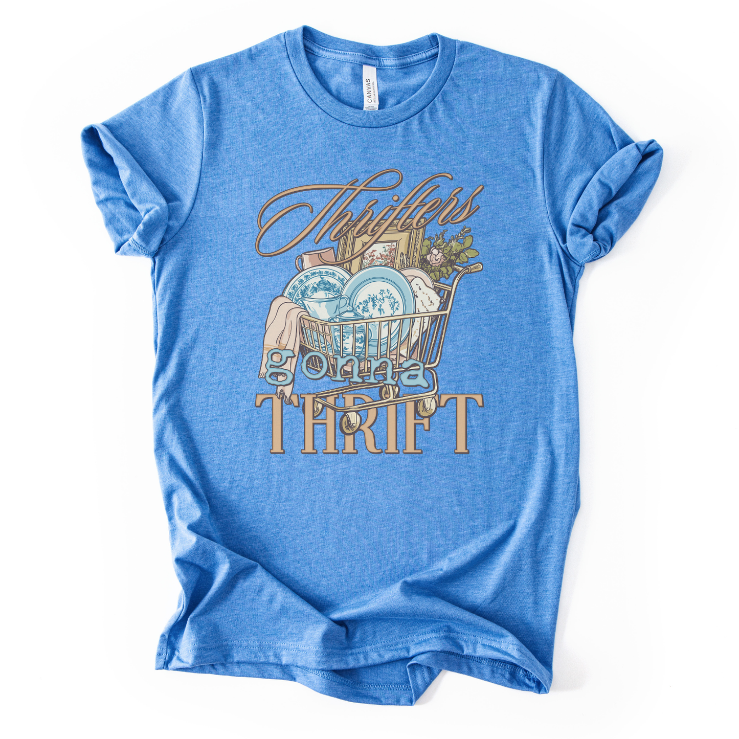 blue t-shirt with graphic design and text on a white background.The trending graphic features an&nbsp;illustrated shopping cart filled with thrifted home decor finds, including blue toile-style plates, folded linens, framed artwork, and greenery. The phrase “Thrifters Gonna Thrift” appears in layered script and bold lettering, creating a warm, lifestyle-driven design that celebrates thrifting culture