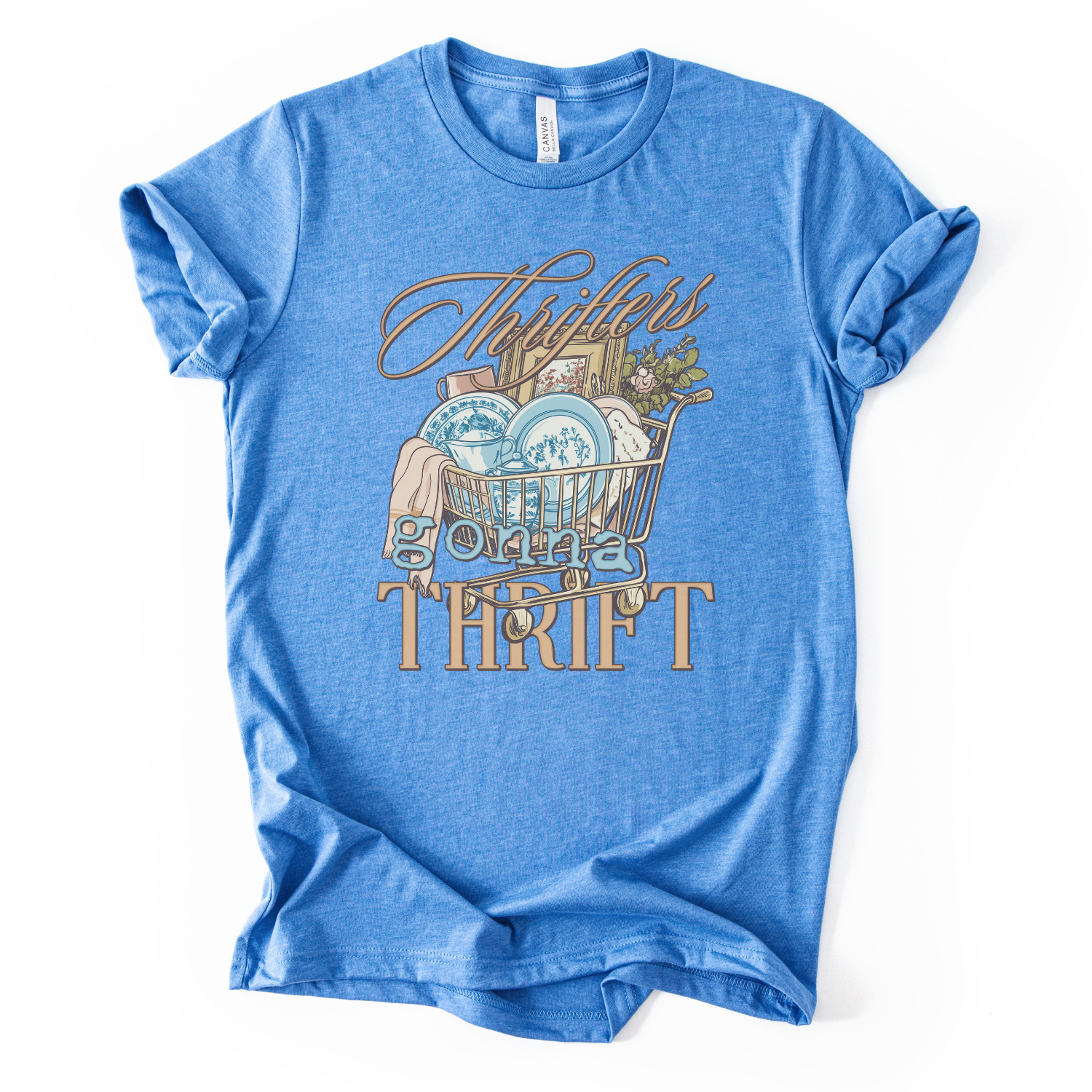 blue t-shirt with graphic design and text on a white background.The trending graphic features an&nbsp;illustrated shopping cart filled with thrifted home decor finds, including blue toile-style plates, folded linens, framed artwork, and greenery. The phrase “Thrifters Gonna Thrift” appears in layered script and bold lettering, creating a warm, lifestyle-driven design that celebrates thrifting culture