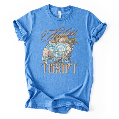blue t-shirt with graphic design and text on a white background.The trending graphic features an&nbsp;illustrated shopping cart filled with thrifted home decor finds, including blue toile-style plates, folded linens, framed artwork, and greenery. The phrase “Thrifters Gonna Thrift” appears in layered script and bold lettering, creating a warm, lifestyle-driven design that celebrates thrifting culture