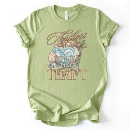 green t-shirt with graphic design and text on a white background.The trending graphic features an&nbsp;illustrated shopping cart filled with thrifted home decor finds, including blue toile-style plates, folded linens, framed artwork, and greenery. The phrase “Thrifters Gonna Thrift” appears in layered script and bold lettering, creating a warm, lifestyle-driven design that celebrates thrifting culture