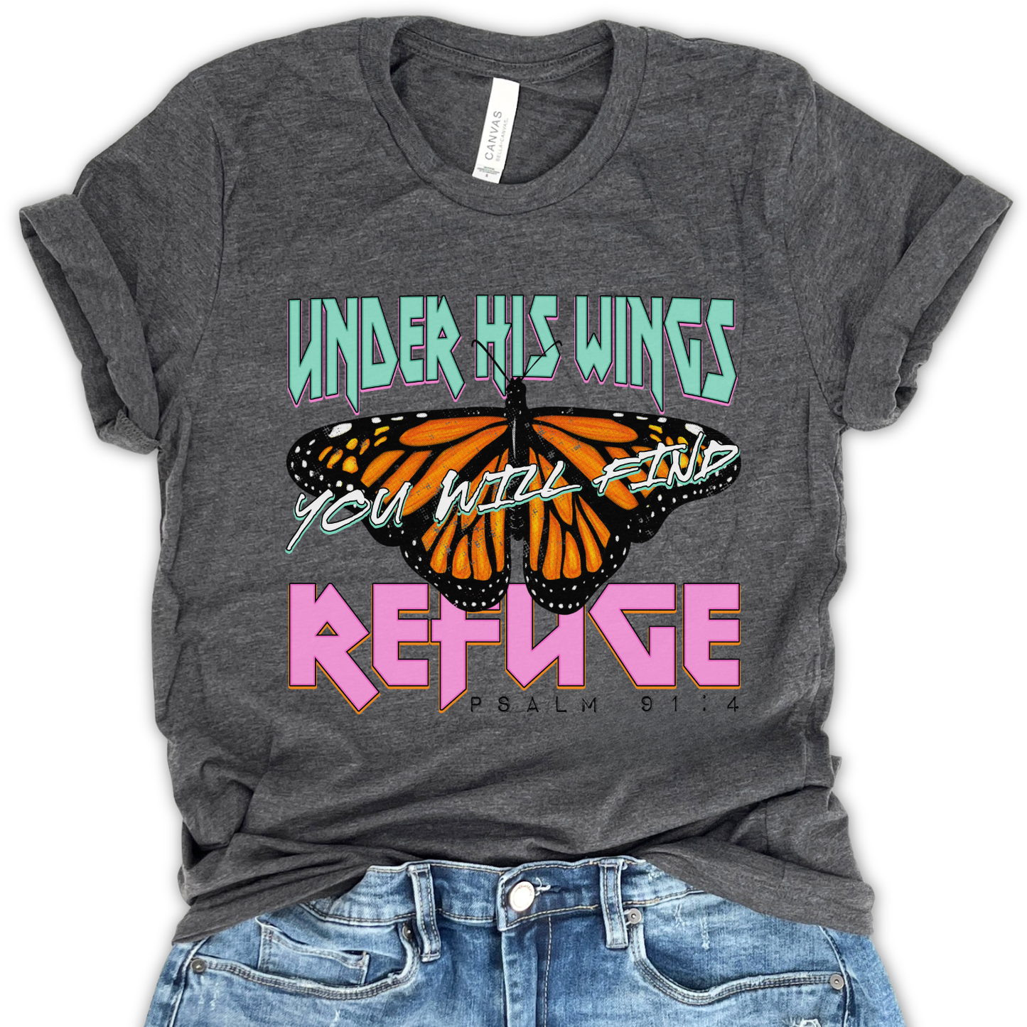 Flat lay of a Bella Canvas unisex graphic t-shirt featuring a monarch butterfly design with bold lettering reading “Under His Wings You Will Find Refuge” and Psalm 91:4, styled casually with denim for a modern faith-inspired look.