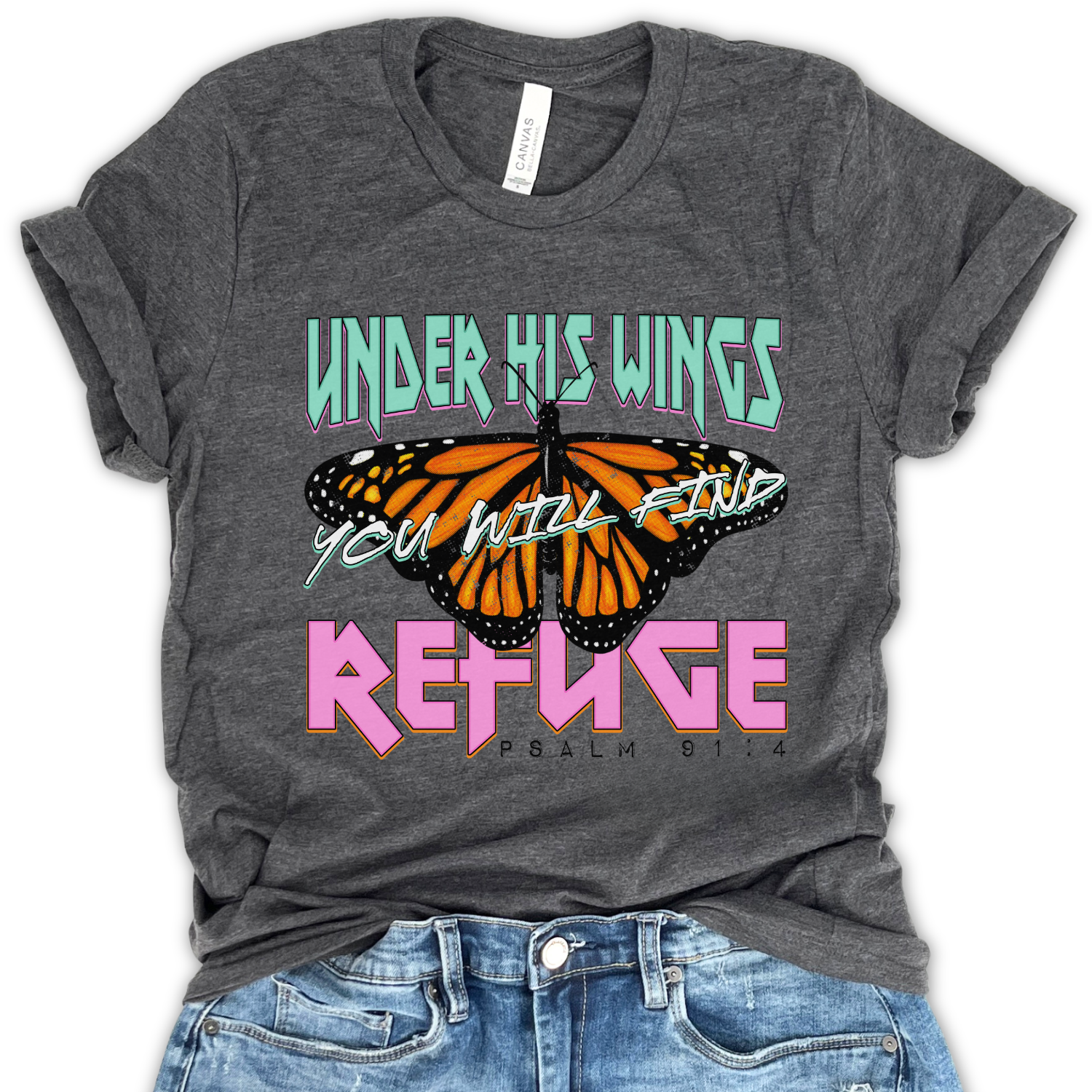 Flat lay of a Bella Canvas unisex graphic t-shirt featuring a monarch butterfly design with bold lettering reading “Under His Wings You Will Find Refuge” and Psalm 91:4, styled casually with denim for a modern faith-inspired look.