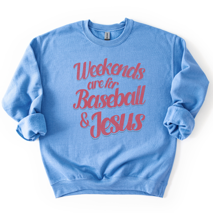 blue-colored crewneck sweatshirt with pink and blue retro script text reading “Weekends are for Baseball & Jesus,” styled flat with sleeves scrunched