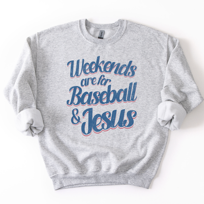 ash-colored crewneck sweatshirt with pink and blue retro script text reading “Weekends are for Baseball & Jesus,” styled flat with sleeves scrunched