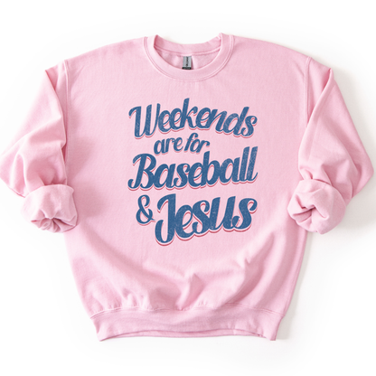 pink-colored crewneck sweatshirt with pink and blue retro script text reading “Weekends are for Baseball & Jesus,” styled flat with sleeves scrunched