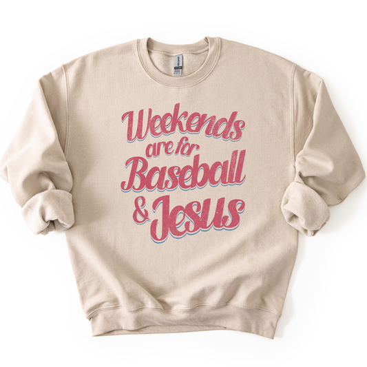Sand-colored crewneck sweatshirt with pink and blue retro script text reading “Weekends are for Baseball & Jesus,” styled flat with sleeves scrunched