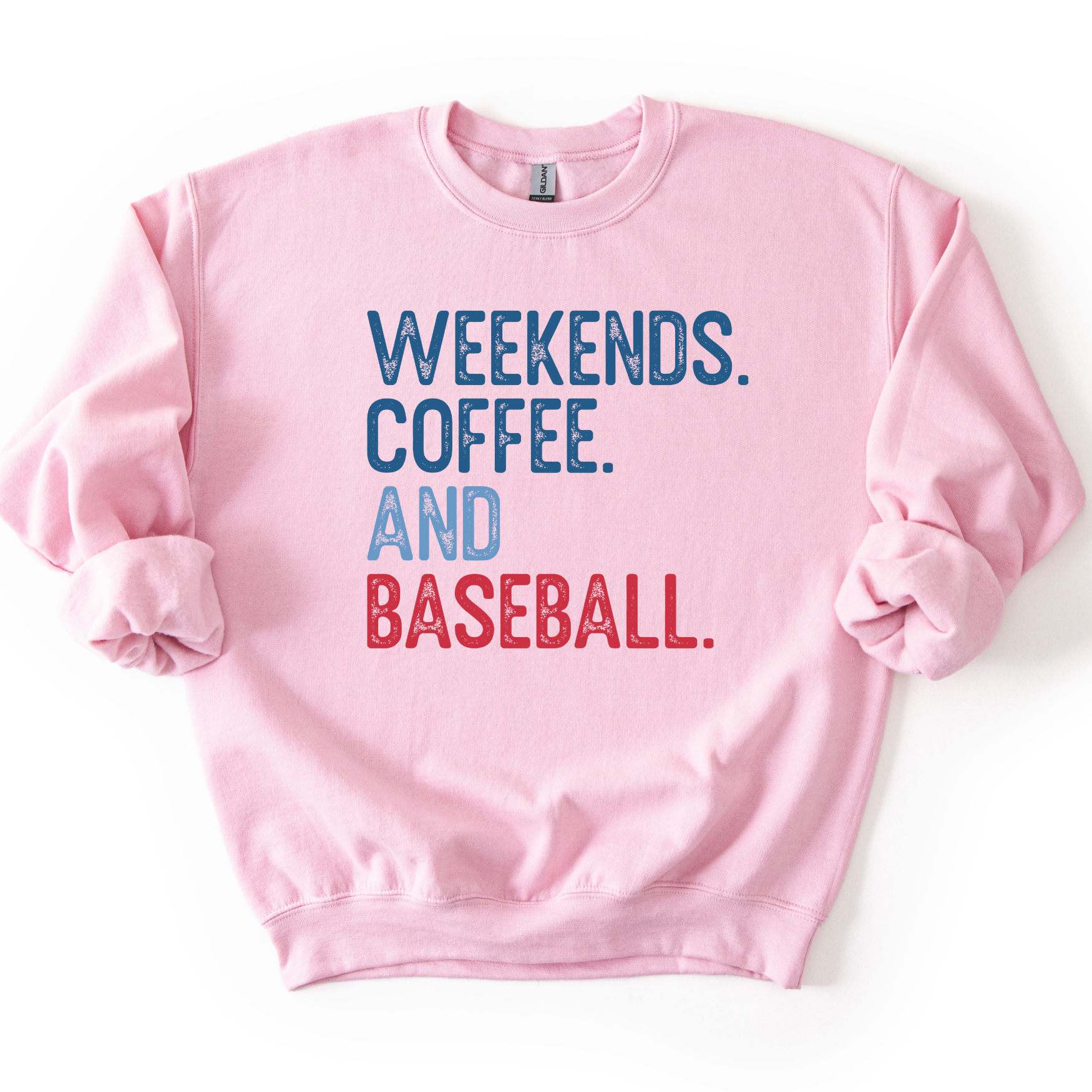 Light pink crewneck sweatshirt with distressed text reading “Weekends. Coffee. And Baseball.” in blue and red lettering, laid flat with sleeves slightly scrunched.