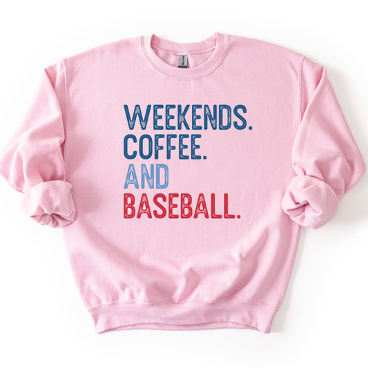 Light pink crewneck sweatshirt with distressed text reading “Weekends. Coffee. And Baseball.” in blue and red lettering, laid flat with sleeves slightly scrunched.